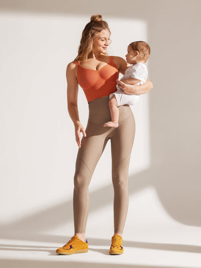 Easelyn Postpartum Recovery Compression Leggings in Tan, full-body front view of a mom holding her baby on her hip